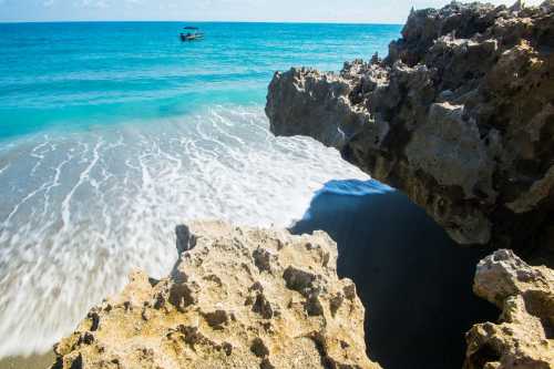 A rocky shoreline with waves gently lapping at the beach, and a small boat in the distance on a clear blue sea.