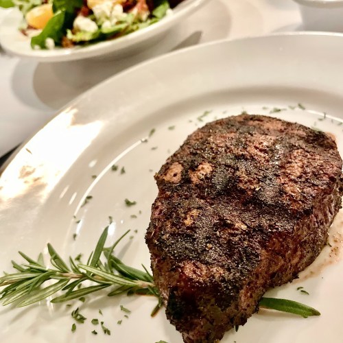 A perfectly cooked steak on a white plate, garnished with rosemary, with a salad in the background.