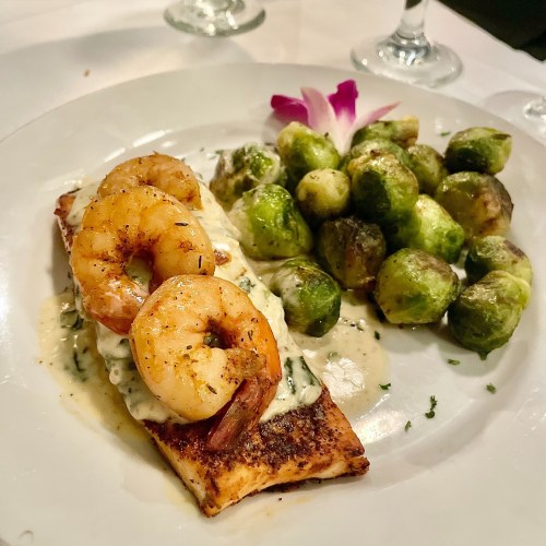 A plate featuring salmon topped with shrimp in a creamy sauce, accompanied by roasted Brussels sprouts and a flower garnish.