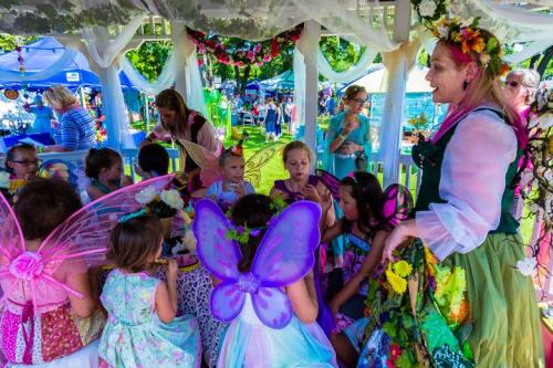 A group of children in colorful fairy costumes gather around a table, enjoying treats at an outdoor event.