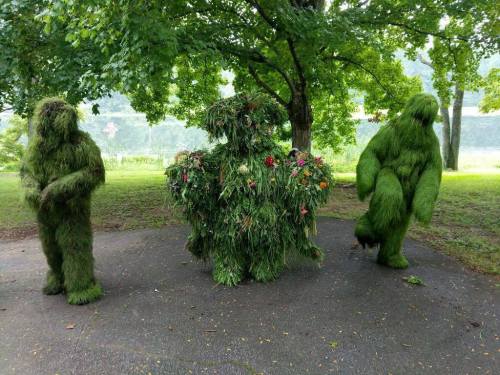 Three figures covered in grass and flowers stand in a park, blending into the lush greenery around them.