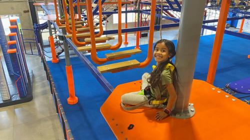 A smiling girl sits on an orange platform in a colorful indoor playground, ready for an adventure on the obstacle course.
