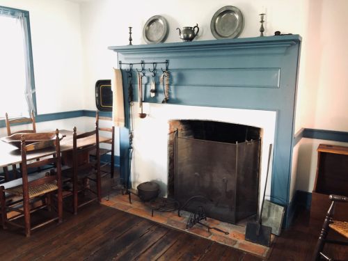 A cozy room featuring a blue fireplace, wooden dining table, chairs, and vintage kitchenware on display.