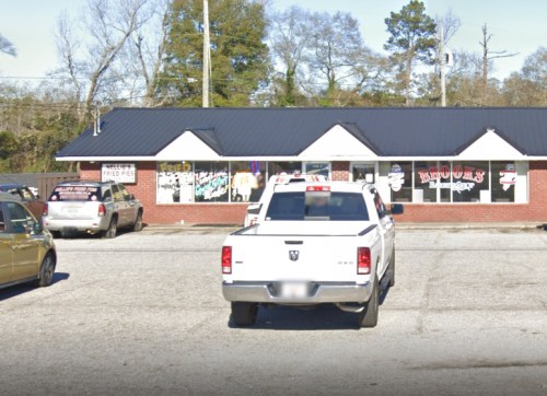 A white pickup truck parked in front of a brick building with colorful signs and a black roof. Trees in the background.