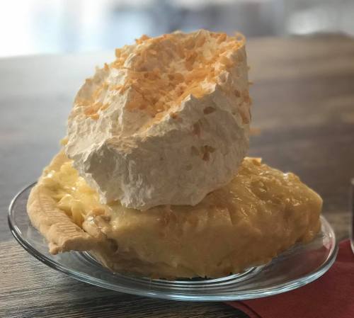 A slice of pie topped with a large mound of whipped cream and toasted coconut on a glass plate.