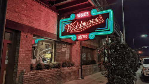 Neon sign for Weldmann's restaurant, established in 1870, glowing at night against a brick building.