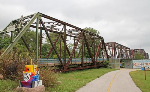 Rusty metal bridge over a path, surrounded by greenery, with a colorful Route 66 sign in the foreground.