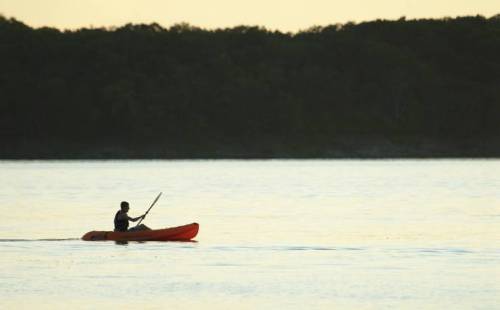 A person paddles a bright orange kayak on calm water, with a forested shoreline in the background at sunset.