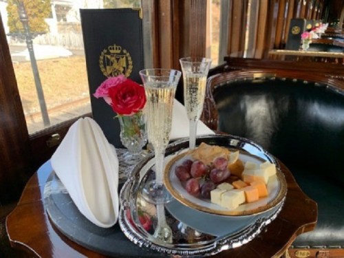 A silver tray with cheese, grapes, and a pastry, accompanied by two champagne flutes and a vase of roses.