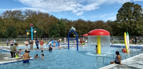 A busy outdoor pool with families enjoying water slides, splash features, and sunny weather. Trees in the background.