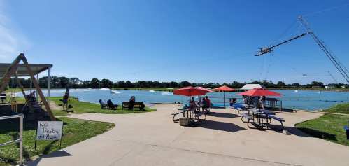 A sunny lakeside scene with picnic tables, umbrellas, and a cable system for water sports in the background.