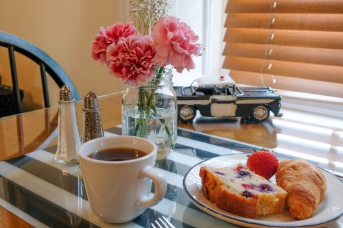 A cozy breakfast scene with coffee, a slice of berry cake, a croissant, and pink flowers in a vase on a table.