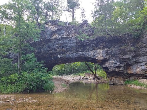 A natural stone arch over a shallow stream, surrounded by lush greenery and trees.