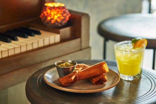 A plate of tamales with a side of green sauce, next to a cocktail and a lit candle on a wooden table by a piano.