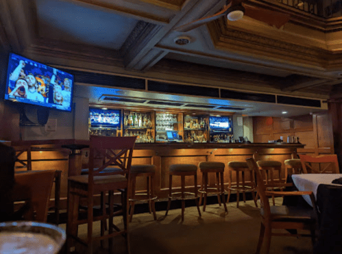 A dimly lit bar area with a wooden counter, high stools, and two TVs displaying sports. Tables are set nearby.