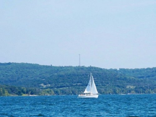 A sailboat glides across a calm blue lake with green hills in the background under a clear sky.