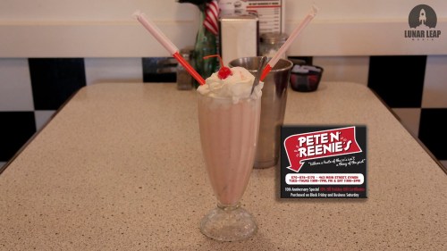 A tall milkshake topped with whipped cream and cherries, served in a retro diner setting with a menu sign nearby.