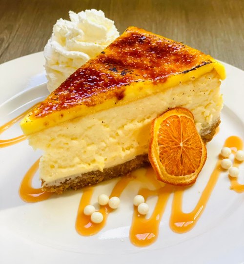 A slice of creamy cheesecake topped with caramelized orange, whipped cream, and decorative sauce on a white plate.