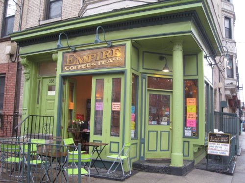 A vibrant green coffee shop with large windows, outdoor seating, and colorful signs advertising drinks and specials.