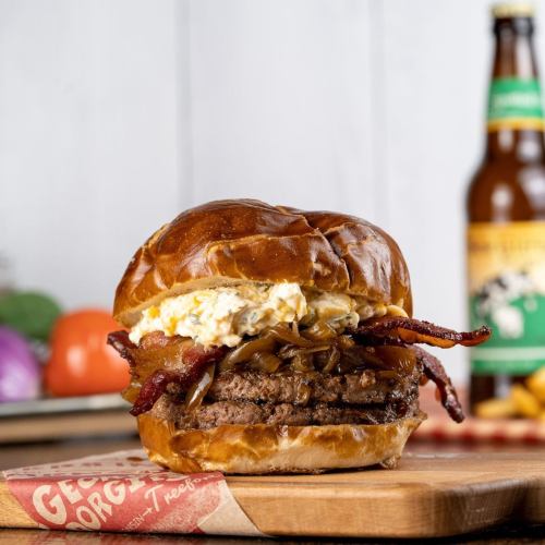 A delicious burger with bacon, caramelized onions, and a creamy topping, served on a pretzel bun with a drink in the background.