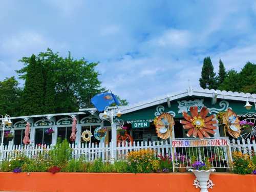 A colorful restaurant with a white picket fence, flowers, and an "Open for Carryout & Dine-In" sign under a blue sky.
