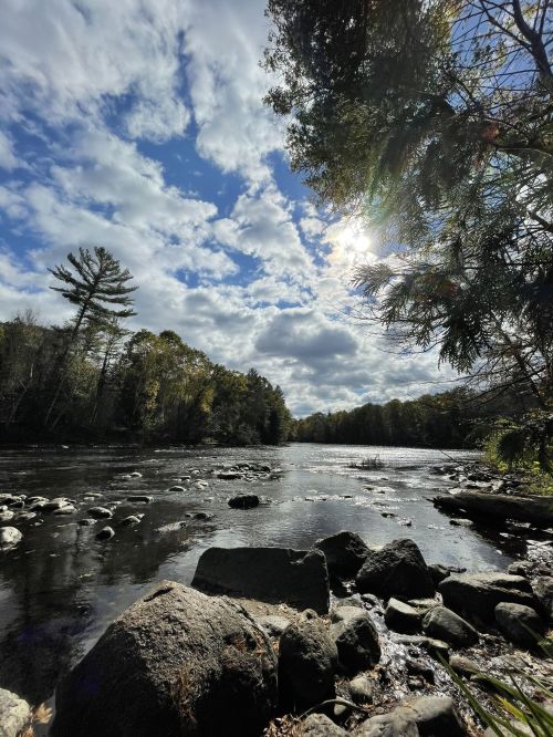 A serene river scene with rocks, trees, and a cloudy sky, illuminated by sunlight peeking through the clouds.