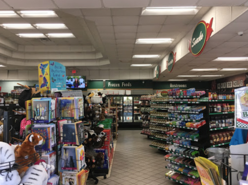 Interior of a grocery store aisle with shelves of snacks, toys, and a frozen foods section in the background.