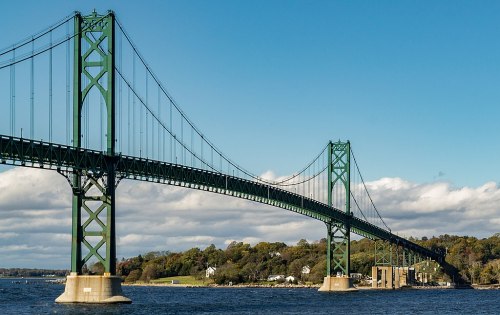 A green suspension bridge spans a river under a clear blue sky, with trees and buildings visible in the background.