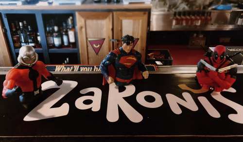 Three superhero figurines—Deadpool, Superman, and another character—displayed on a bar counter with the name "Zaronis."