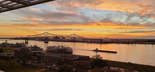 A vibrant sunset over a river, with a bridge in the background and a barge passing by.