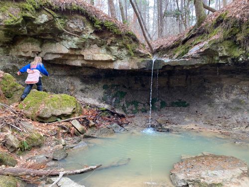 A child explores a rocky area with a small waterfall and pool, surrounded by moss and trees in a forest setting.