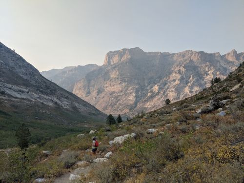 A runner traverses a rocky trail in a valley surrounded by towering mountains and a hazy sky.