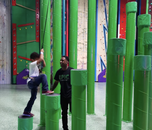 A child climbs green cylindrical structures while an adult watches in a colorful indoor play area.