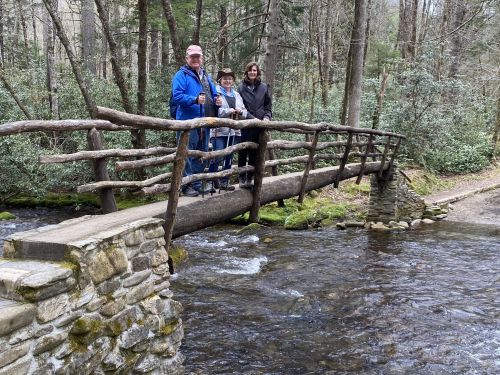 Three people stand on a rustic wooden bridge over a stream, surrounded by trees and greenery.