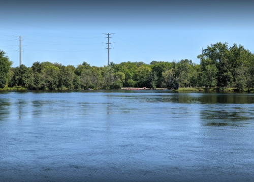 A calm river scene with trees lining the shore and power lines in the background under a clear blue sky.
