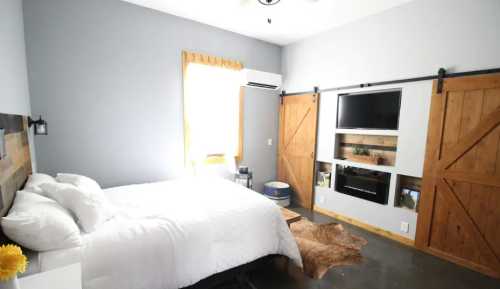 Cozy bedroom with a white bed, wooden accents, a TV, and sliding barn doors, featuring natural light from a window.