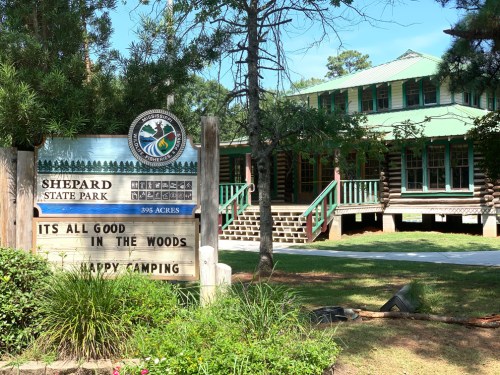 Sign for Shepard State Park with a log cabin in the background, surrounded by trees and a clear blue sky.