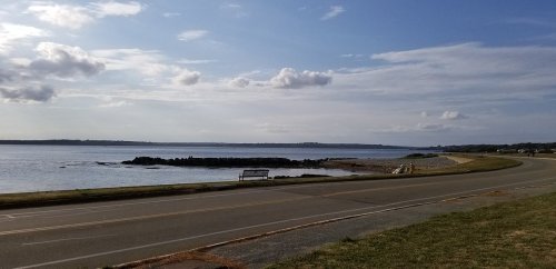 A serene coastal view with a calm sea, distant land, and a road lined with grass under a partly cloudy sky.