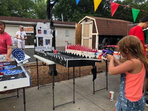A child aims a toy gun at a carnival game, while adults enjoy nearby attractions decorated with colorful flags.
