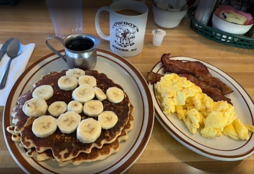 A plate of pancakes topped with banana slices, scrambled eggs, crispy bacon, and a cup of coffee on a diner table.