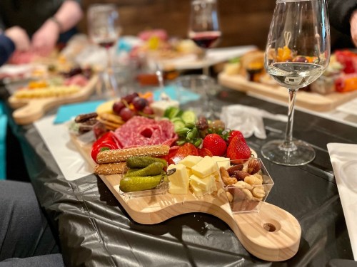 A wooden platter with assorted meats, cheeses, fruits, nuts, and pickles, accompanied by wine glasses on a table.