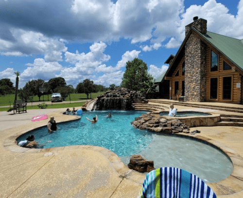 A sunny pool scene with people relaxing in and around a stone pool, featuring a waterfall and a log cabin in the background.