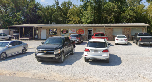A small brick building with a sign reading "Clay's" and several parked cars in a gravel lot. Trees in the background.