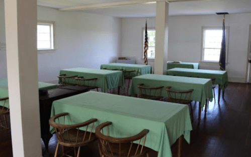 A spacious room with several tables covered in green cloth and wooden chairs, featuring windows and flags.