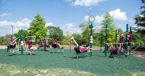 Children in maroon shirts play on a green playground with various fitness equipment under a blue sky.