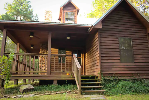 A rustic wooden cabin with a porch, surrounded by trees and greenery, featuring a stone chimney and a gabled roof.