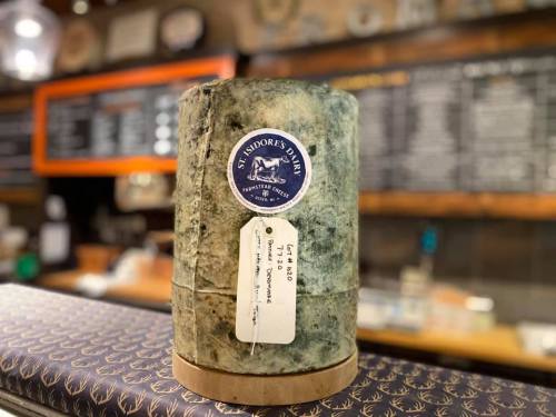 A round cheese with a blue-green rind, labeled "St. Adore's Blue," displayed on a wooden board in a rustic setting.
