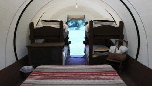 Interior of a cozy, rustic cabin with bunk beds, a double bed, and a small table, illuminated by a hanging light.