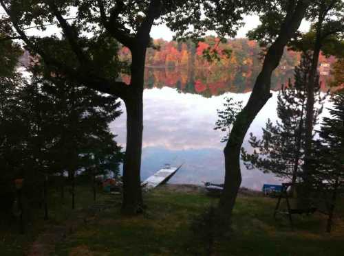 A serene lake scene framed by trees, reflecting autumn colors in the water. Boats are visible along the shore.