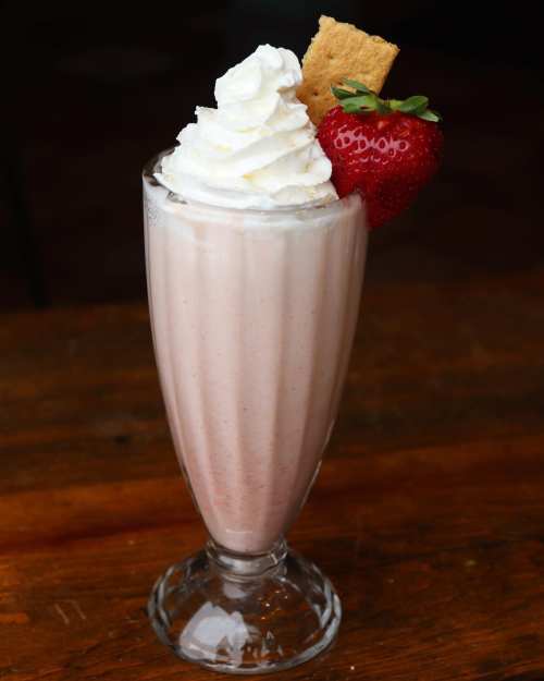 A tall glass of strawberry milkshake topped with whipped cream, a fresh strawberry, and a graham cracker.